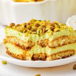 A delectable slice of pistachio tiramisu graces a white plate, boasting layers of rich cream and delicate ladyfingers, all crowned with a sprinkle of chopped pistachios.