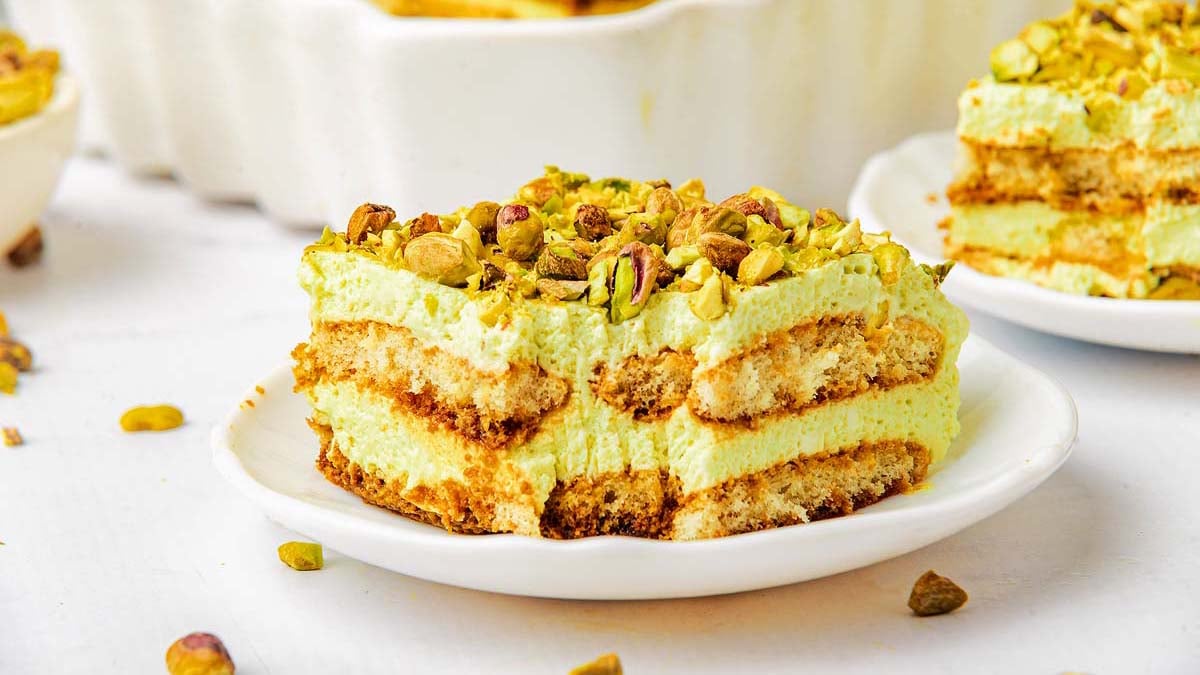 A slice of pistachio tiramisu topped with chopped pistachios on a white plate. Layers of cream and sponge cake are visible.