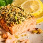 Close-up of pistachio-crusted salmon fillets topped with a herb and nut crust, served with lemon slices on a plate and garnished with fresh spinach leaves.