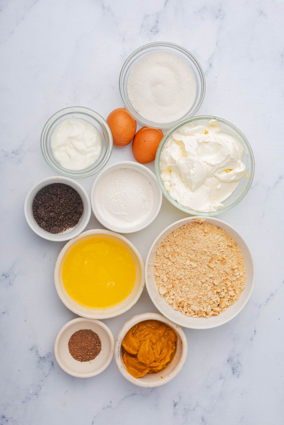 Various ingredients, such as eggs, sugar, crushed crackers, yogurt, cream, melted butter, flour, poppy seeds, and spices are artfully arranged on a marble surface ready to be transformed into delicious pumpkin cheesecake bars.