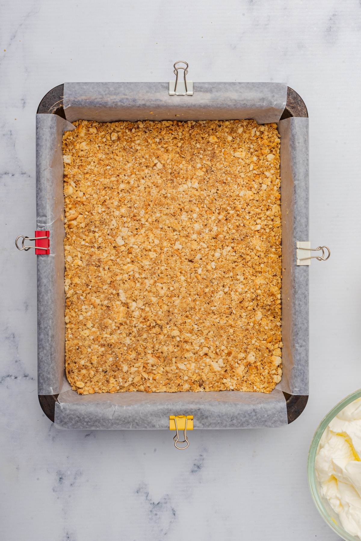 A square baking pan with parchment paper clipped to the sides cradles a crumbly, golden crust that's the perfect foundation for decadent pumpkin cheesecake bars.