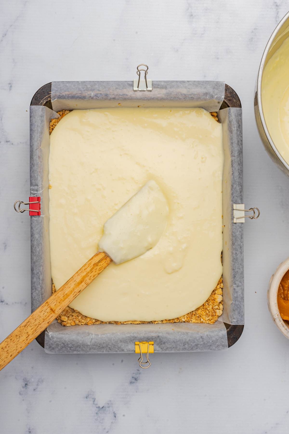 Square baking pan with parchment paper, filled with creamy pumpkin cheesecake batter being spread by a wooden spatula. Mixing bowl and small cup are beside the pan.
