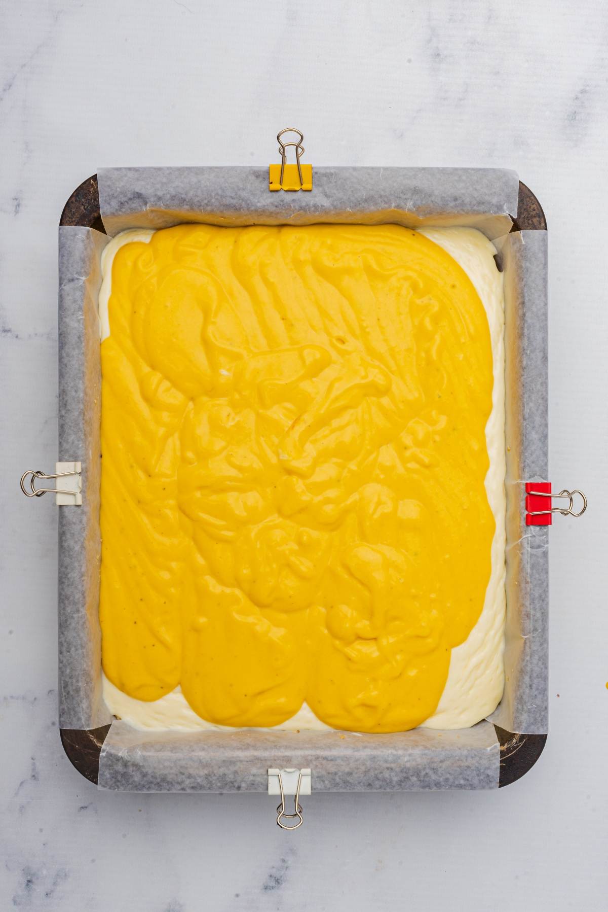 Pumpkin cheesecake bars in a baking pan feature two layers of batter, one white and one yellow, secured with paper clips on a marbled surface.
