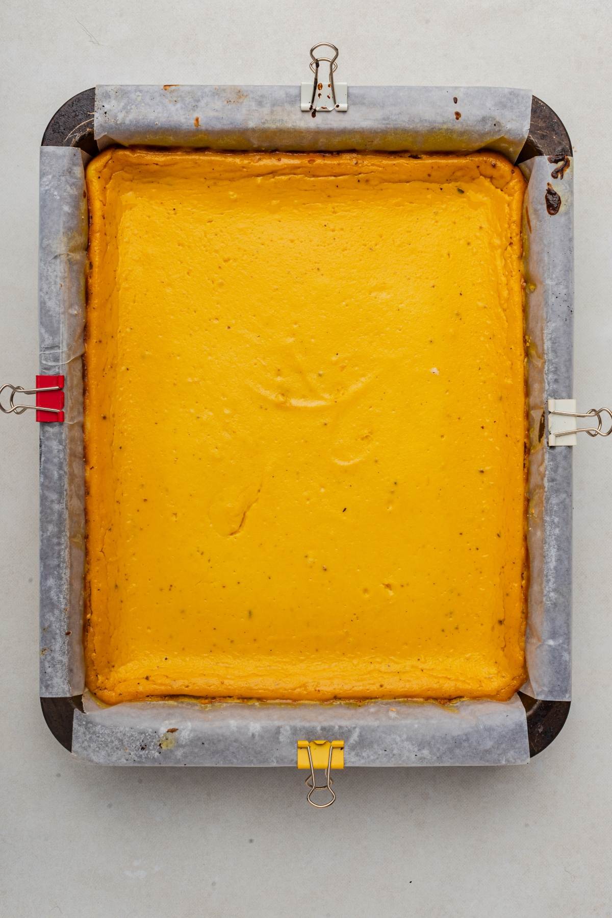 Square orange pumpkin cheesecake bars rest in a parchment-lined baking tray, secured with metal clips.