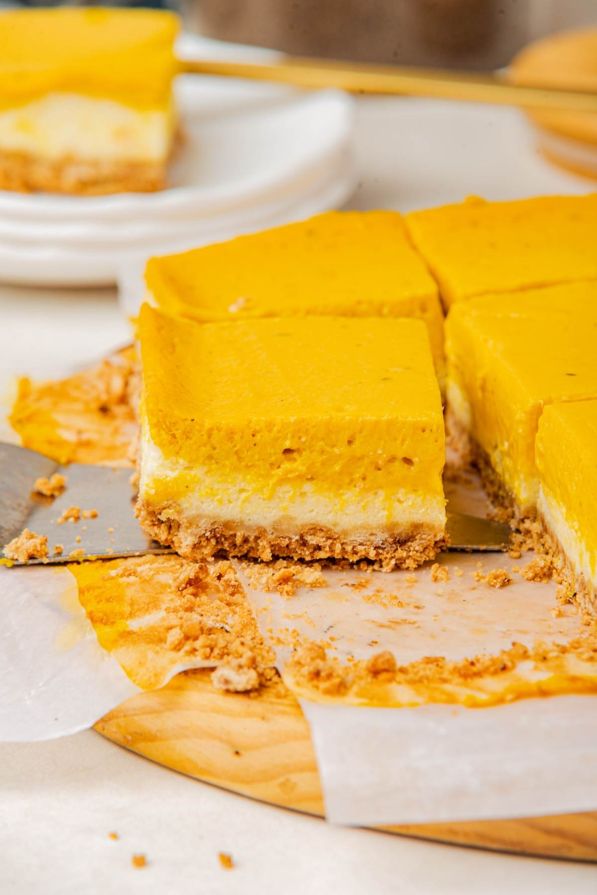 A sliced pumpkin cheesecake with a yellow topping rests on a wooden board, crumbs scattered around. A serving spatula is partially inserted under one piece.
