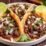 Three tacos filled with slow cooker Mexican shredded beef, chopped onions, and cilantro, served with lime halves on a white plate.