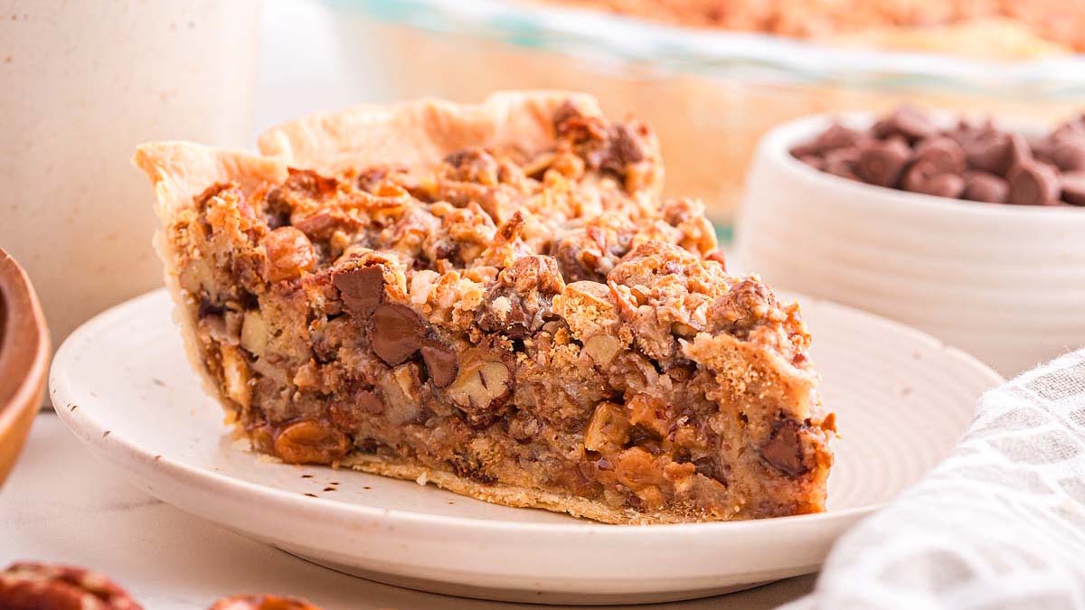 A slice of pie with a crumbly crust filled with pecans and chocolate chips on a white plate.