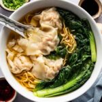 A steaming bowl of wonton noodle soup with greens sits elegantly on a gray surface, encircled by fresh ingredients like ginger, garlic, and an array of condiments.