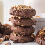 A stack of Texas sheet cake cookies, rich with chocolate and topped with crunchy nuts, is surrounded by two milk bottles and a small white bowl in the background.