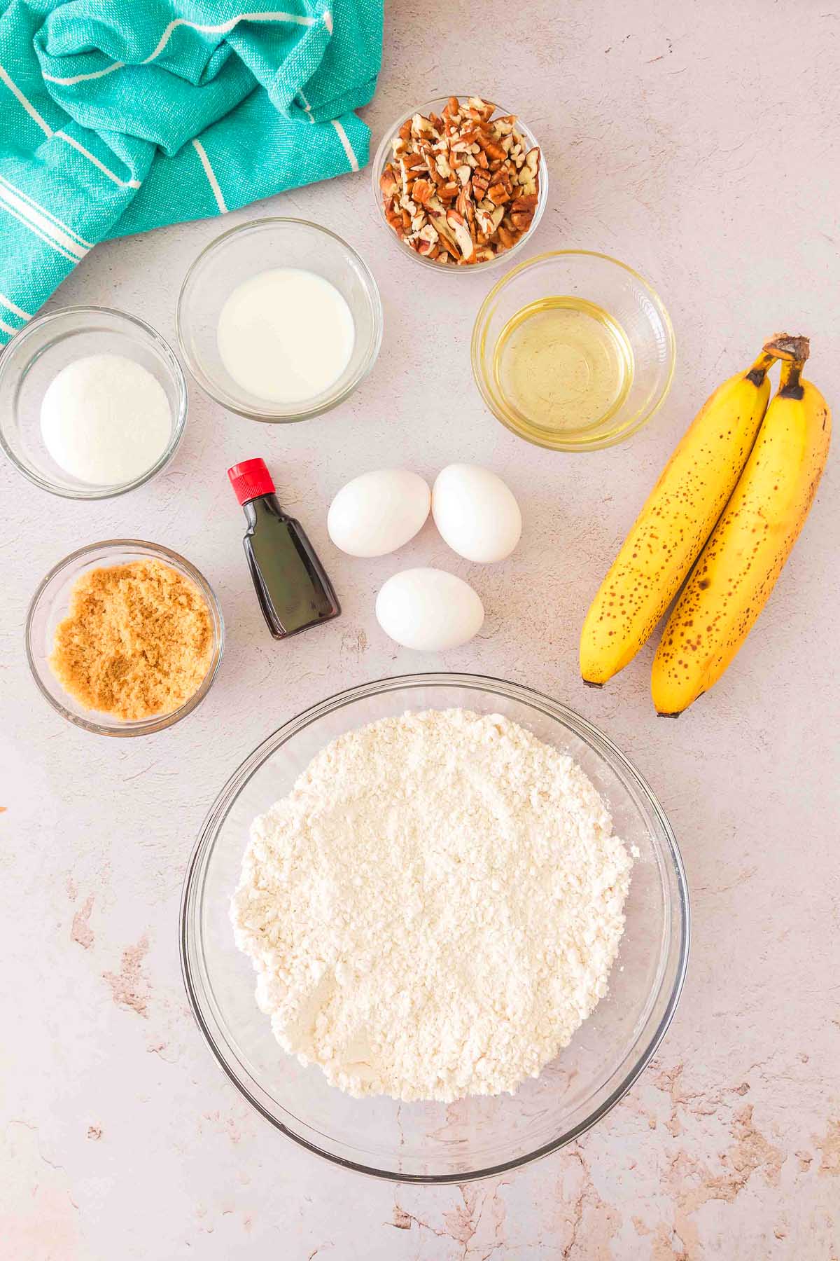 Baking ingredients for Bisquick banana bread, including Bisquick, eggs, bananas, milk, nuts, vanilla, oil, and sugar on a light countertop.