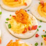Cajun shrimp deviled eggs are adorned with bacon bits and chopped chives, elegantly presented on a white plate.