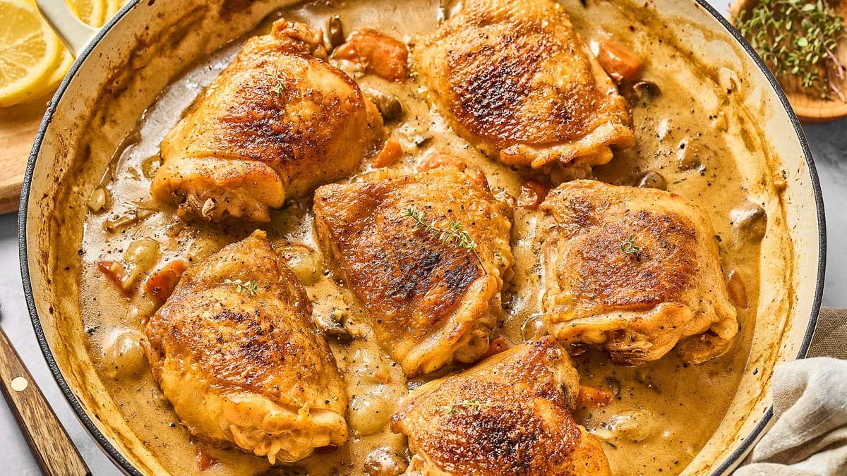 A skillet with browned chicken thighs in a creamy sauce with mushrooms and herbs.