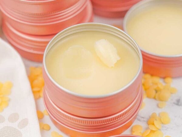 Round metal tins filled with creamy Dog Paw Balm, with yellow wax pellets scattered on a white surface.