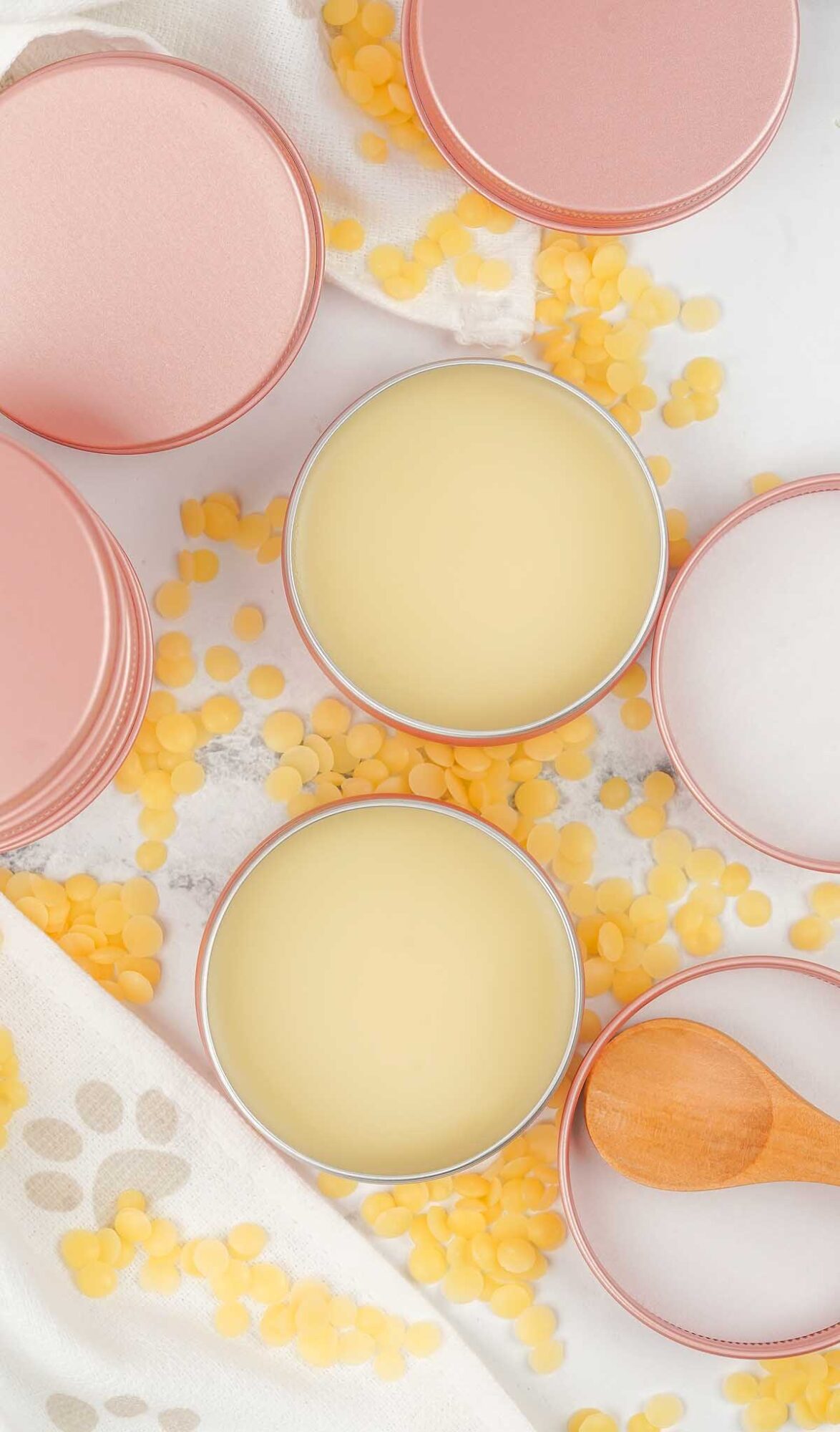 Open tins of Dog Paw Balm with pink lids, yellow wax pellets, and a small wooden spoon are arranged on a white surface.