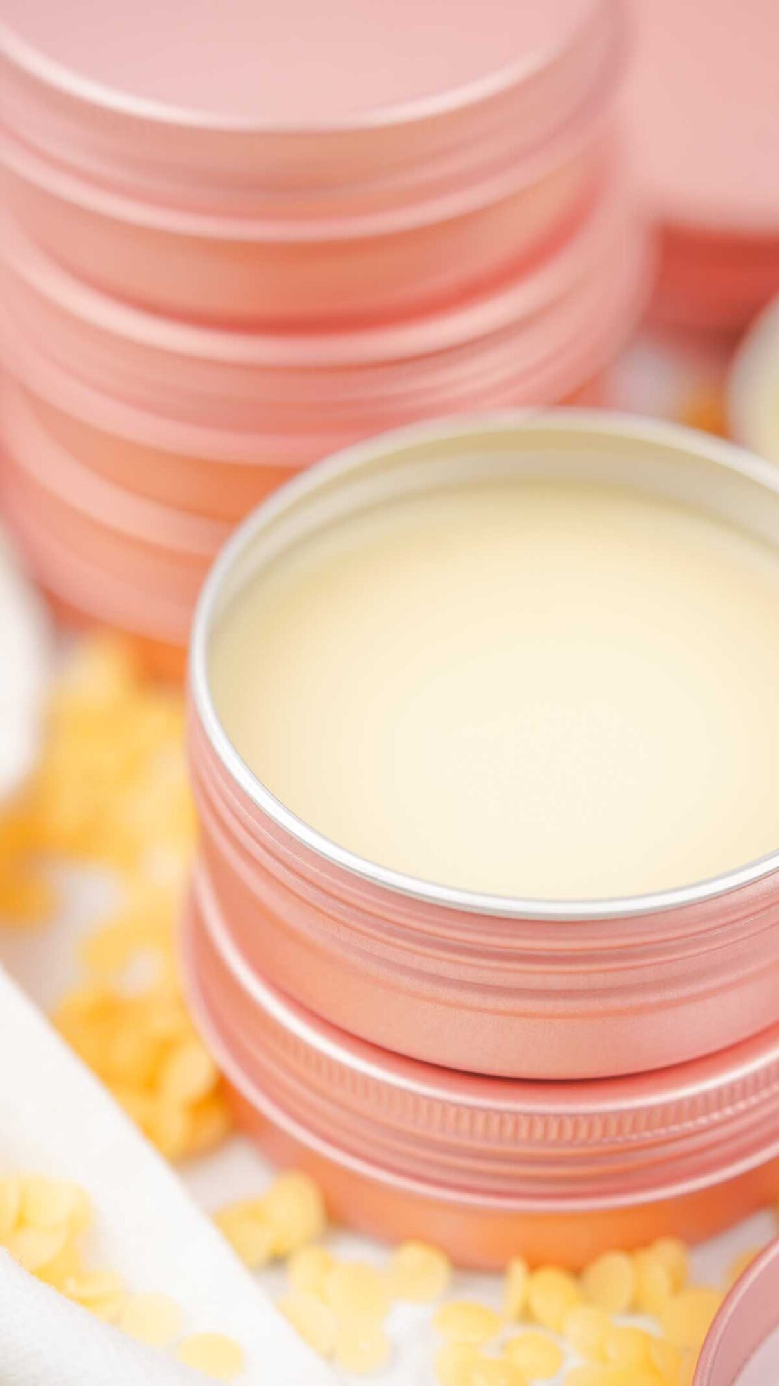 A pink tin of Dog Paw Balm sits surrounded by yellow beeswax pellets, with more stacked tins in the background.