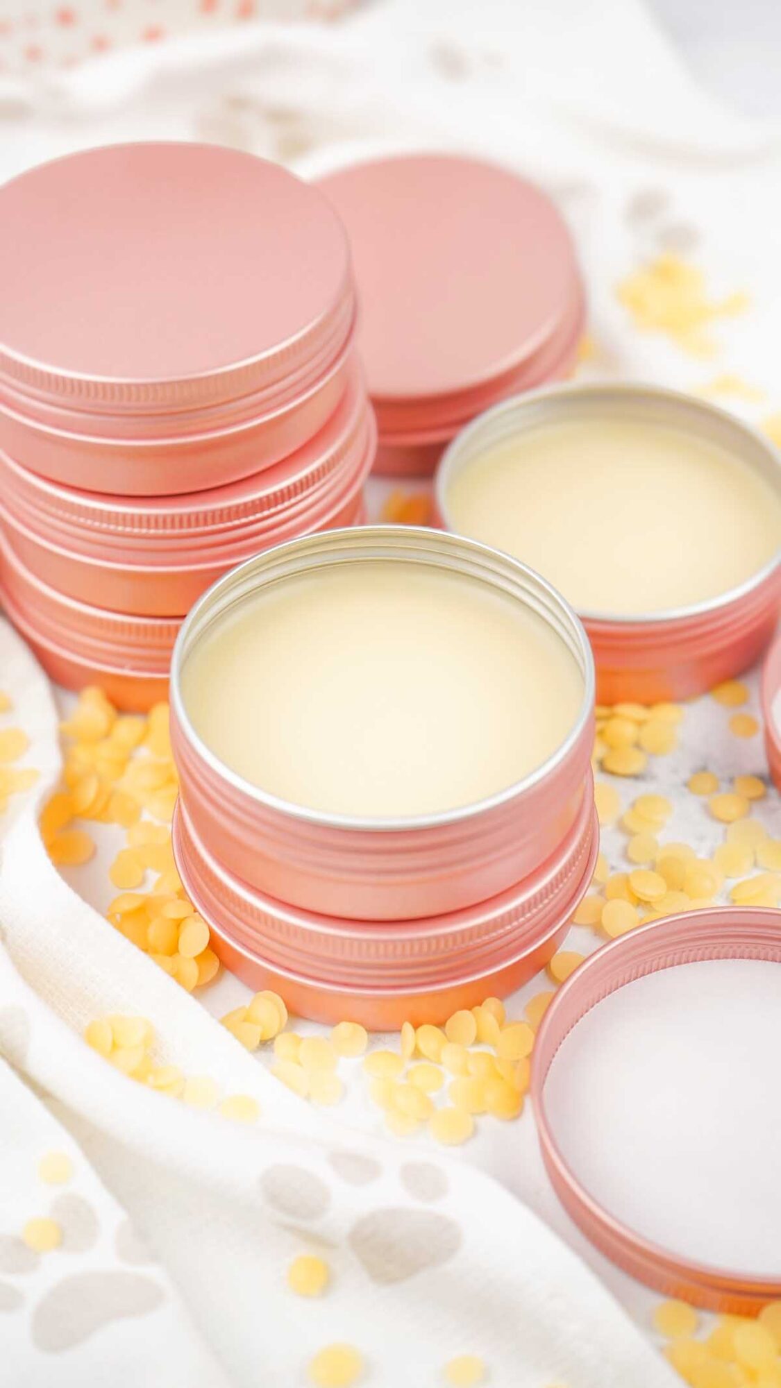Pink tins filled with Dog Paw Balm, surrounded by scattered yellow wax pellets on a light cloth.