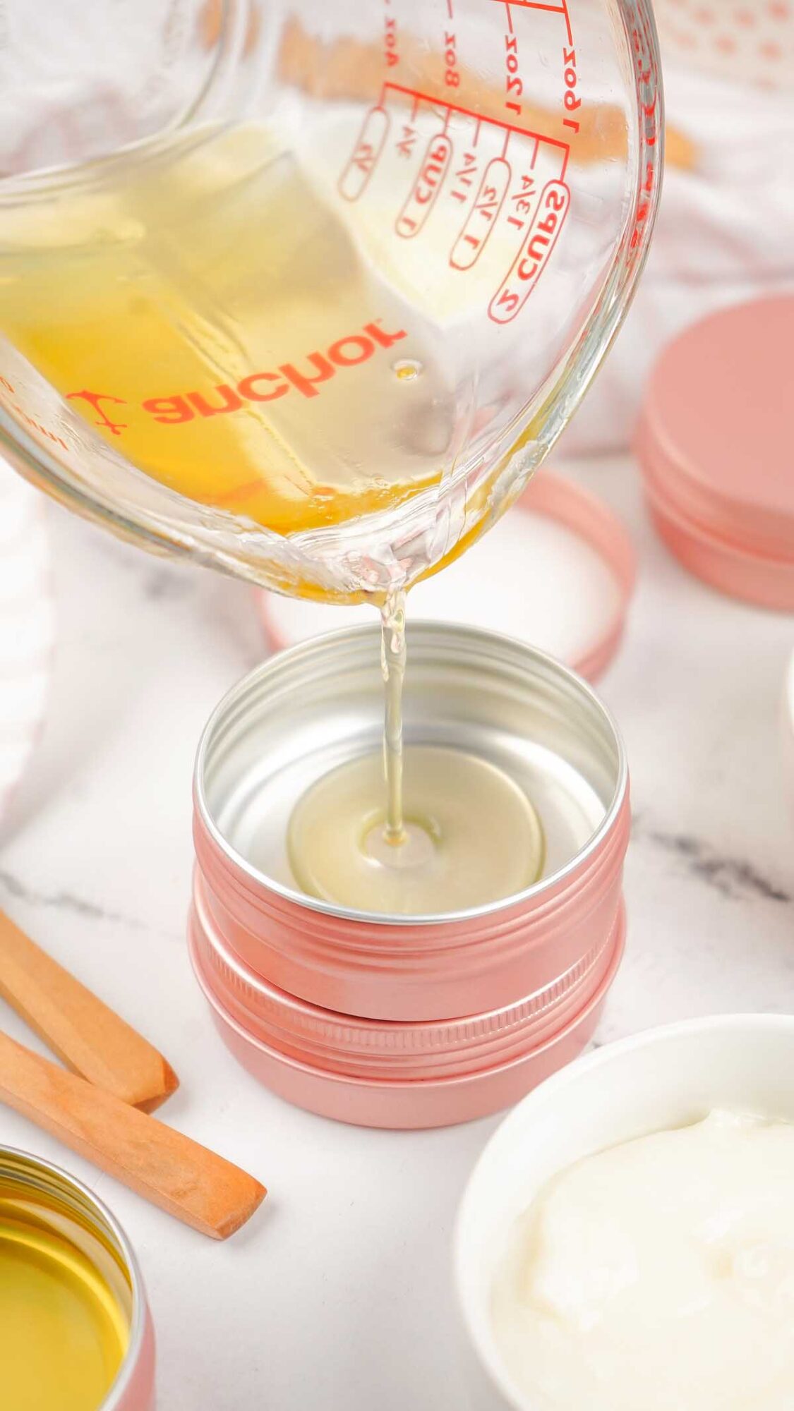 Liquid Dog Paw Balm is being poured from a measuring cup into a small, round metal tin with a pink lid on a white surface.