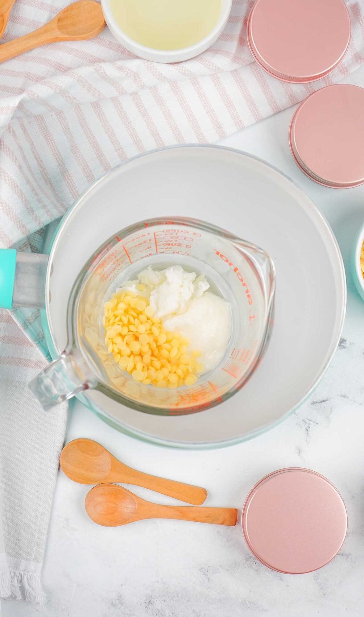 Top view of a measuring cup with solid ingredients for Dog Paw Balm, wooden spoons, pink tins, and a striped towel nearby.