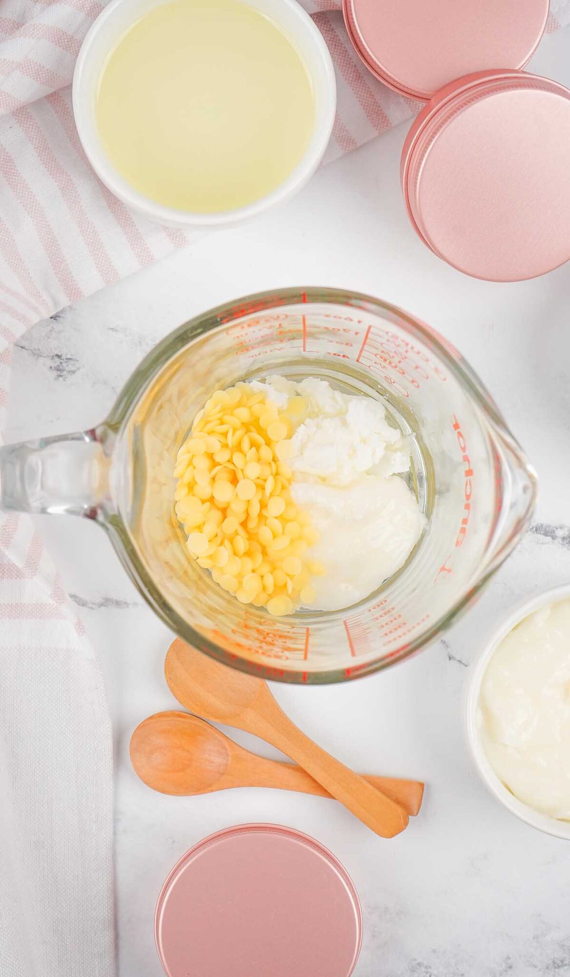 A glass measuring cup with yellow pellets and white cream for Dog Paw Balm, surrounded by pink tins and wooden spoons on a marble surface.