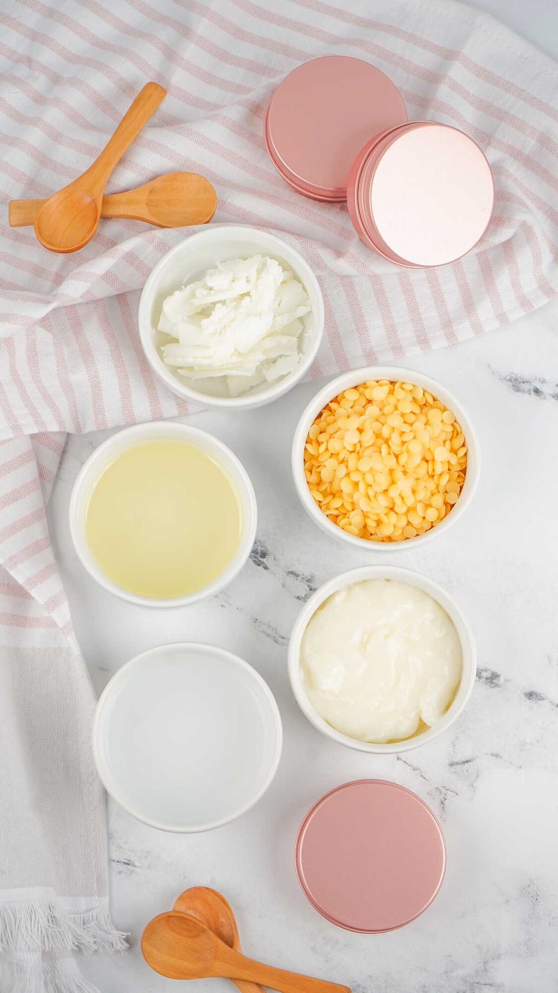 Five bowls with skincare ingredients and pink tins of Dog Paw Balm rest on a striped towel, with wooden spoons on a marble surface.