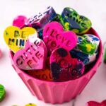 A pink cup brims with colorful DIY heart-shaped crayons, each emblazoned with sweet messages like "I LOVE YOU" and "CUTIE PIE."