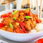 A bowl of rice topped with Kung Pao chicken, red bell peppers, peanuts, and green onions sits invitingly with chopsticks on the side. A napkin, soy sauce, and garlic in the background complete this delightful scene.