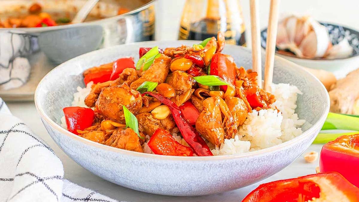 Bowl of stir-fried chicken with red bell peppers, peanuts, and green onions on white rice, next to chopsticks and cooking ingredients.