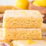 A stack of glazed lemon brownies with vibrant lemon zest on top, surrounded by more tempting bars and fresh whole lemons in the background.