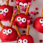 Red cake pops shaped like reindeer with pretzel antlers, candy eyes, and heart-shaped noses rest on a white surface. Love bug cake balls add a touch of sweetness to this whimsical scene, with small heart-shaped sprinkles scattered around.