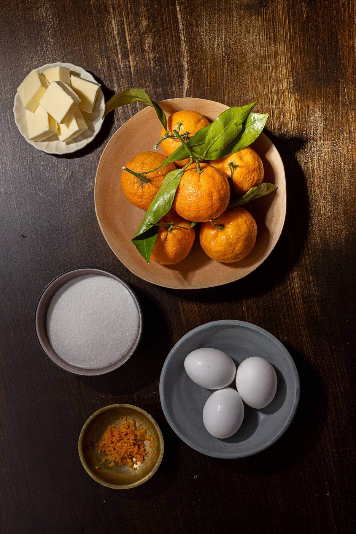 A wooden table is adorned with ingredients: a bowl of mandarins with leaves, three eggs, a plate of butter, a bowl of sugar, and a dish of orange zest—perfect for making mandarin orange curd.