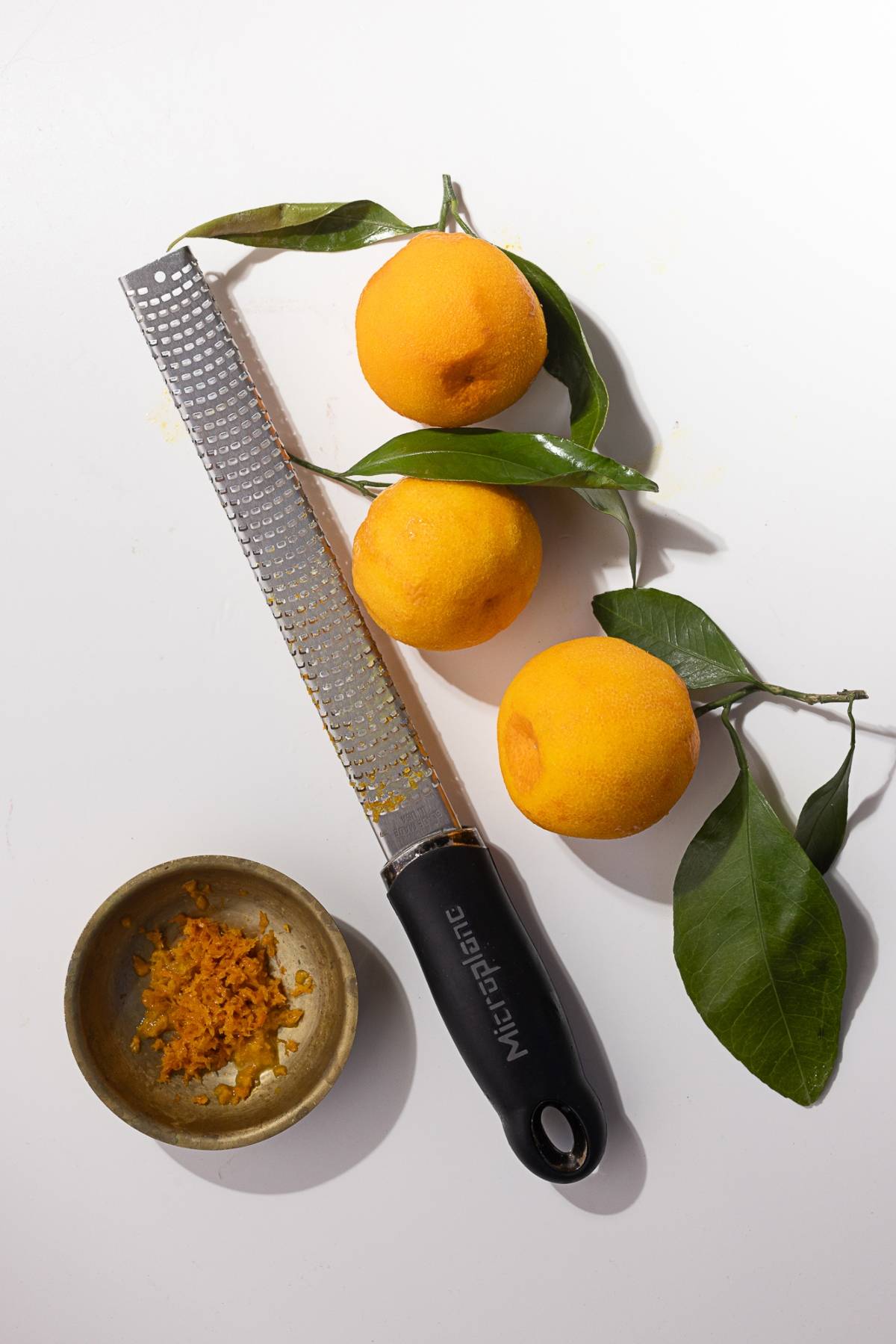 A grater and three vibrant mandarin oranges with leaves rest on a pristine white surface. Nearby, a bowl brims with fresh orange zest, perfect for creating a silky mandarin orange curd.