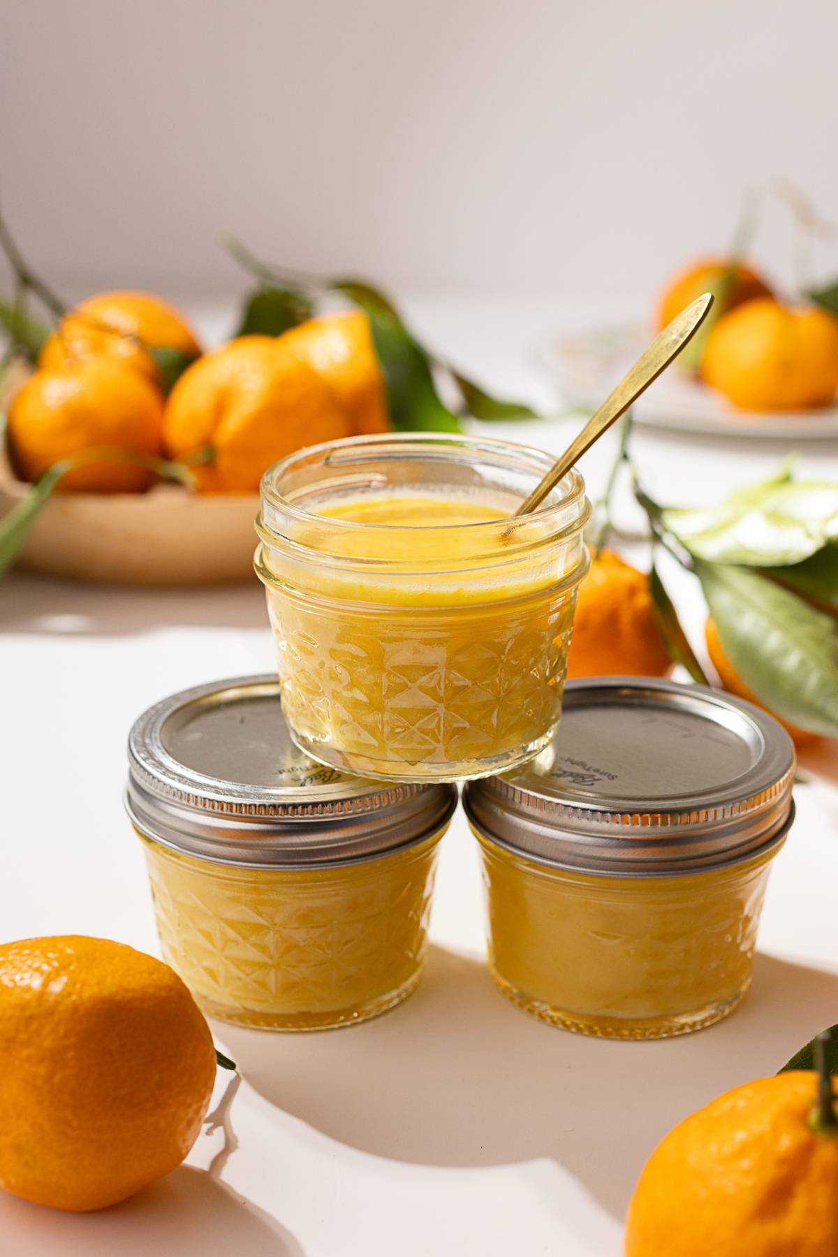 Three jars of lemon curd are artfully arranged on a table, accompanied by fresh oranges and leaves in the background. One jar, filled with a delightful mandarin orange curd, is open with a spoon inside.