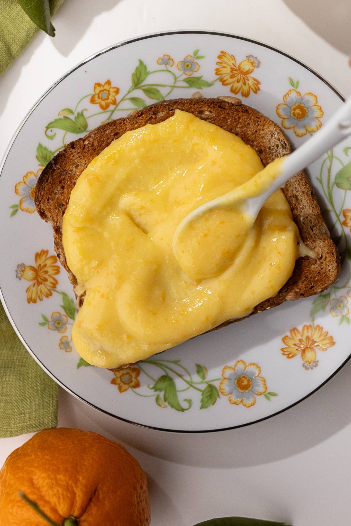 Slice of toast topped with yellow mandarin orange curd on a floral-patterned plate, with a spoon gracefully spreading the topping. A whole orange sits nearby.