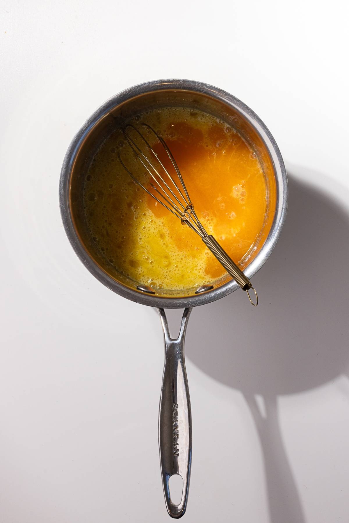 A metal saucepan with beaten eggs and a whisk resting inside, hinting at the creation of a delightful mandarin orange curd, sits on a pristine white surface.
