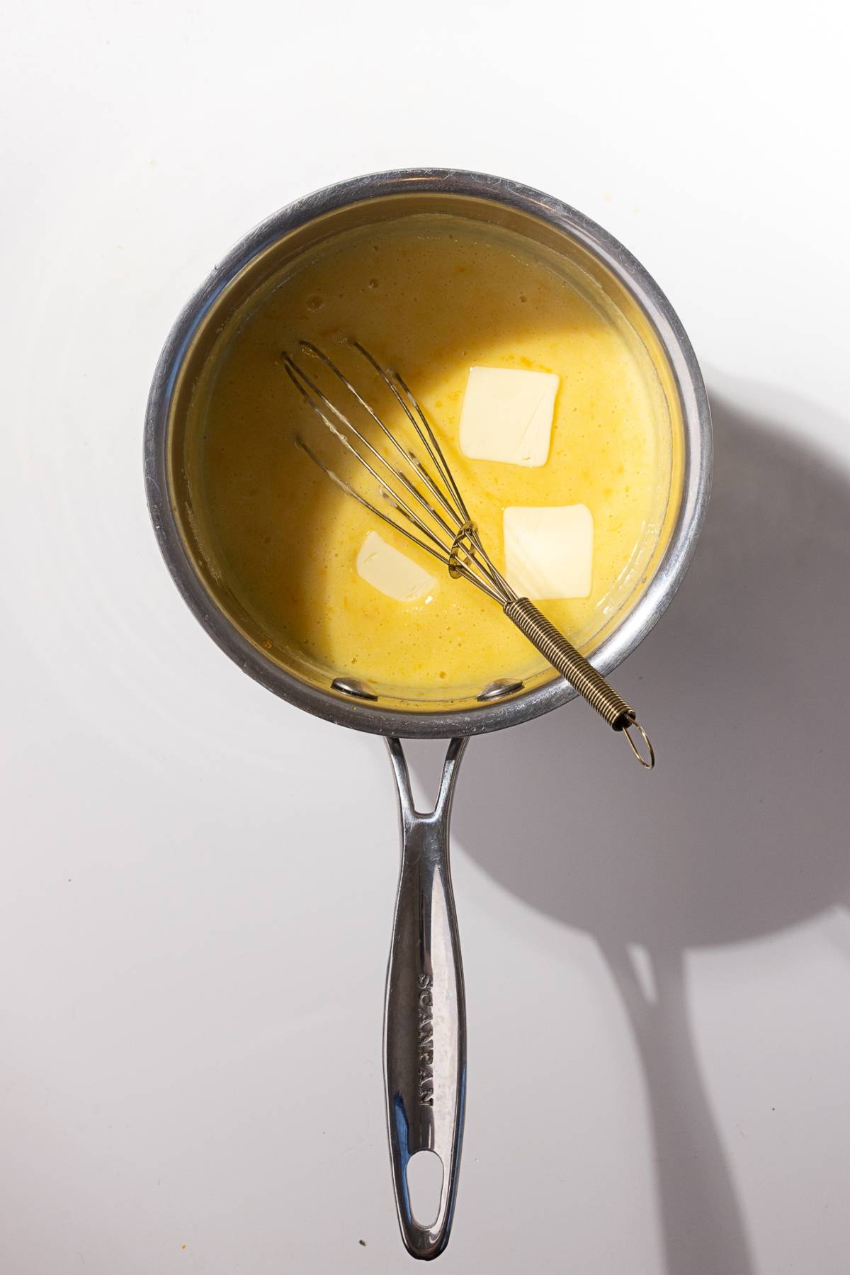 A saucepan holds a creamy yellow mixture, rich and smooth, with two butter slices melting in. The hint of mandarin orange curd adds a zesty twist, while a whisk rests inside, ready to blend the flavors seamlessly.
