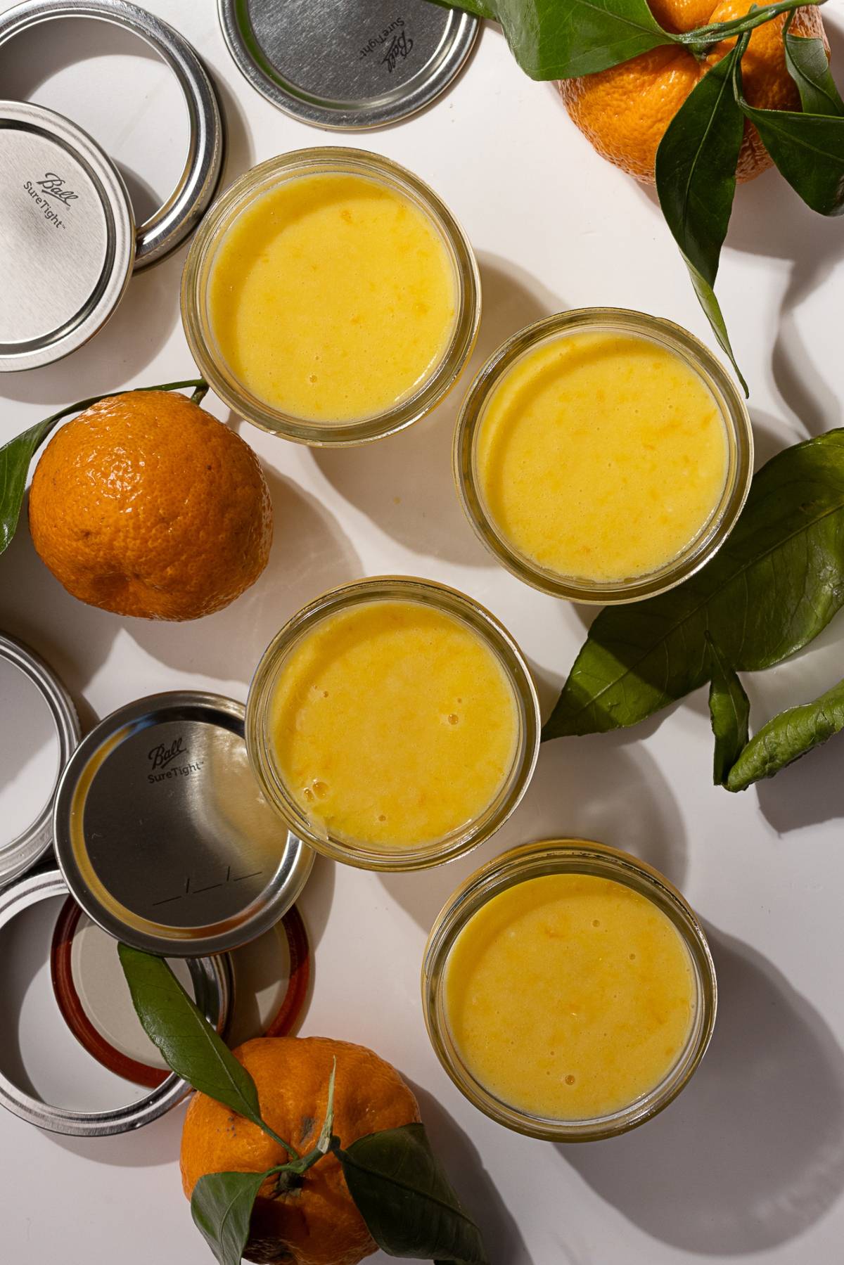 Four glass jars filled with a rich orange smoothie are surrounded by whole mandarin oranges, their vibrant green leaves adding a touch of freshness. Nearby, metal lids lie scattered on the white surface, evoking the look of homemade mandarin orange curd ready to be sealed and savored.