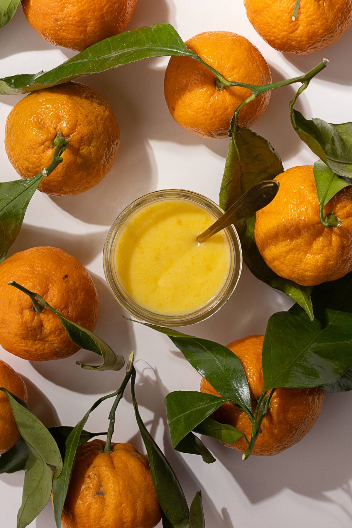 A small glass bowl filled with yellow liquid, resembling mandarin orange curd, is surrounded by whole oranges with green leaves on a white surface.