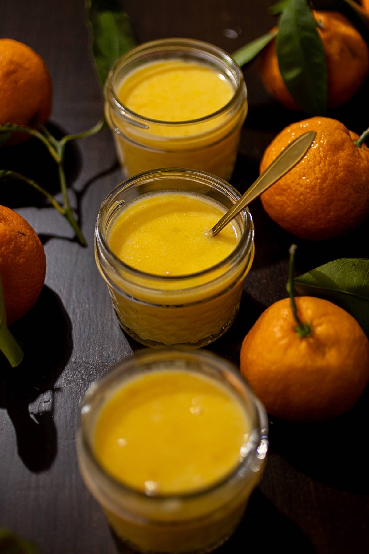Three jars of mandarin orange curd custard with a spoon in the middle jar, surrounded by whole oranges with green leaves on a dark surface.