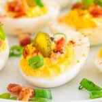 Million dollar deviled eggs topped with crispy bacon, sliced pickles, and fresh green onions are elegantly arranged on a white plate.
