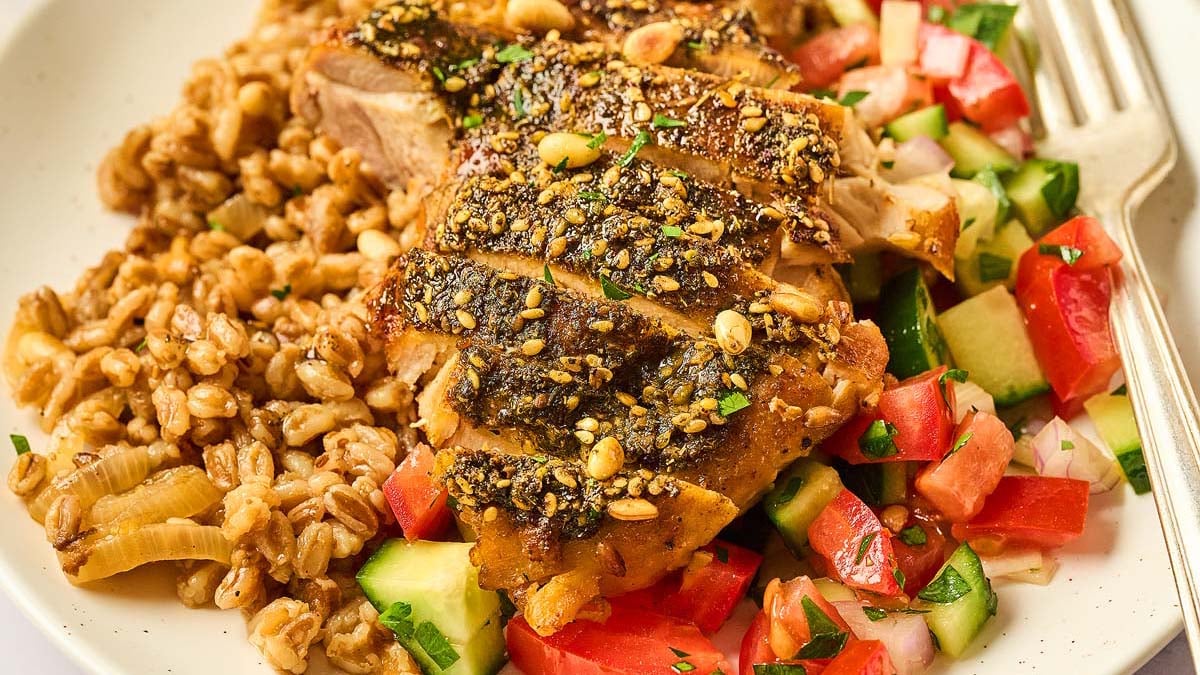 A plate with seasoned grilled chicken, farro, and diced vegetables including tomatoes, cucumbers, and onions, with a fork on the side.