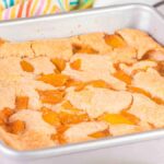 A Bisquick peach cobbler bakes in a square metal pan, its golden-brown crust perfectly enveloping the juicy peach slices. A striped cloth and stacked dishes add to the cozy tableau in the background.