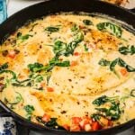A skillet with creamy chicken Florentine featuring tender chicken breasts, spinach, and diced tomatoes in a rich sauce. A wooden spoon, napkin, and glass of water are nearby.