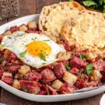 A delicious plate of corned beef hash with a sunny-side-up egg, accompanied by buttered English muffins. A fork rests invitingly on the plate, while salt, pepper, and fresh parsley add to the enticing scene in the background.