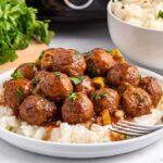 A plate of slow cooker Mississippi meatballs in sauce sits over creamy mashed potatoes, garnished with a sprinkle of parsley. A fork rests beside it, while in the background, a bowl of velvety mashed potatoes and fresh parsley awaits.