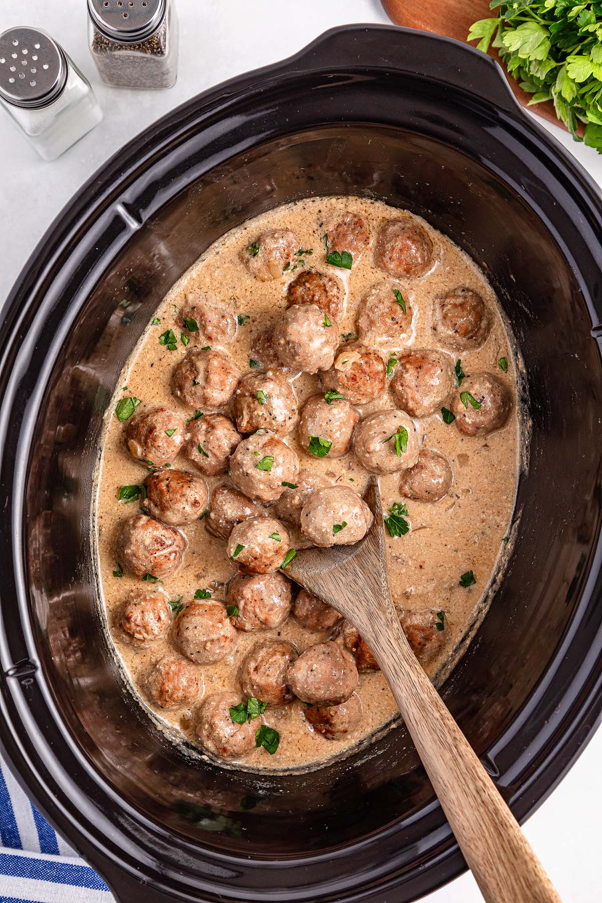 A Crockpot brims with Swedish meatballs in a creamy sauce, garnished with chopped herbs. A wooden spoon stirs the savory blend, while salt and pepper shakers stand ready in the background.