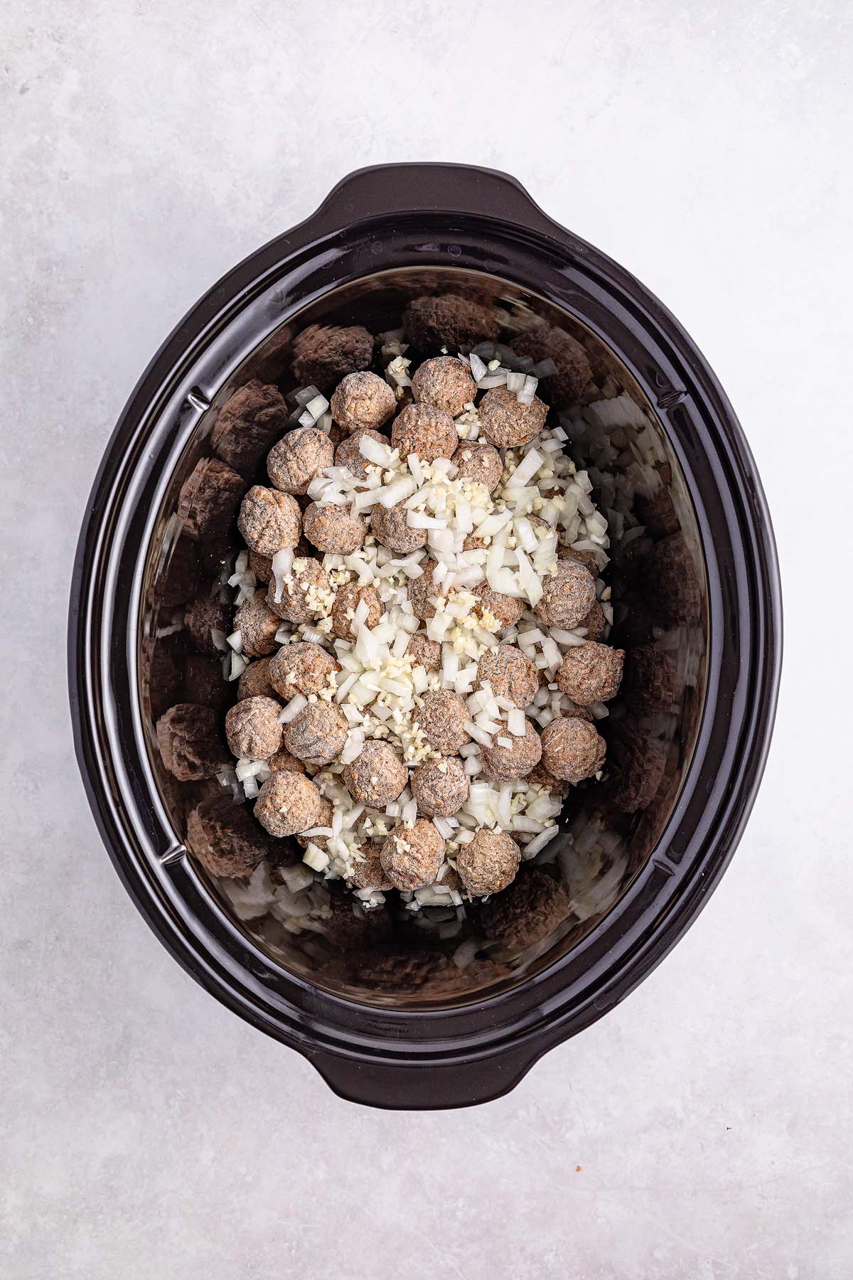 Slow cooker with uncooked Swedish meatballs, chopped onions, and minced garlic, viewed from above.