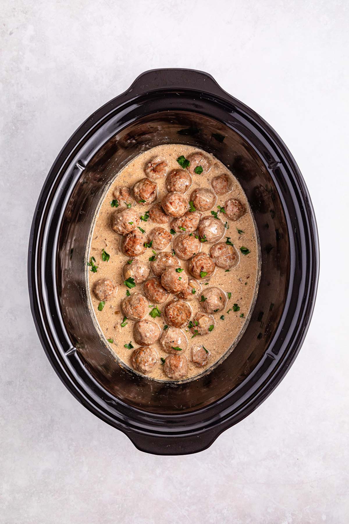 A slow cooker brimming with Swedish meatballs, enveloped in a creamy sauce and garnished with chopped herbs.