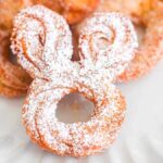 A delightful bunny churro, perfectly shaped like a bunny face and dusted with powdered sugar, rests on a pristine white plate.