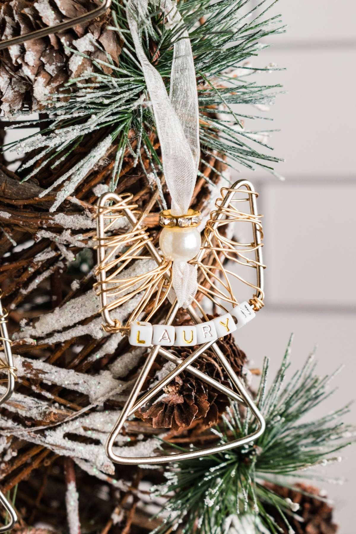 A gold wire angel ornament with pearl head and white ribbon hangs on a frosted evergreen branch, with the name "LAURYN" spelled in beads.
