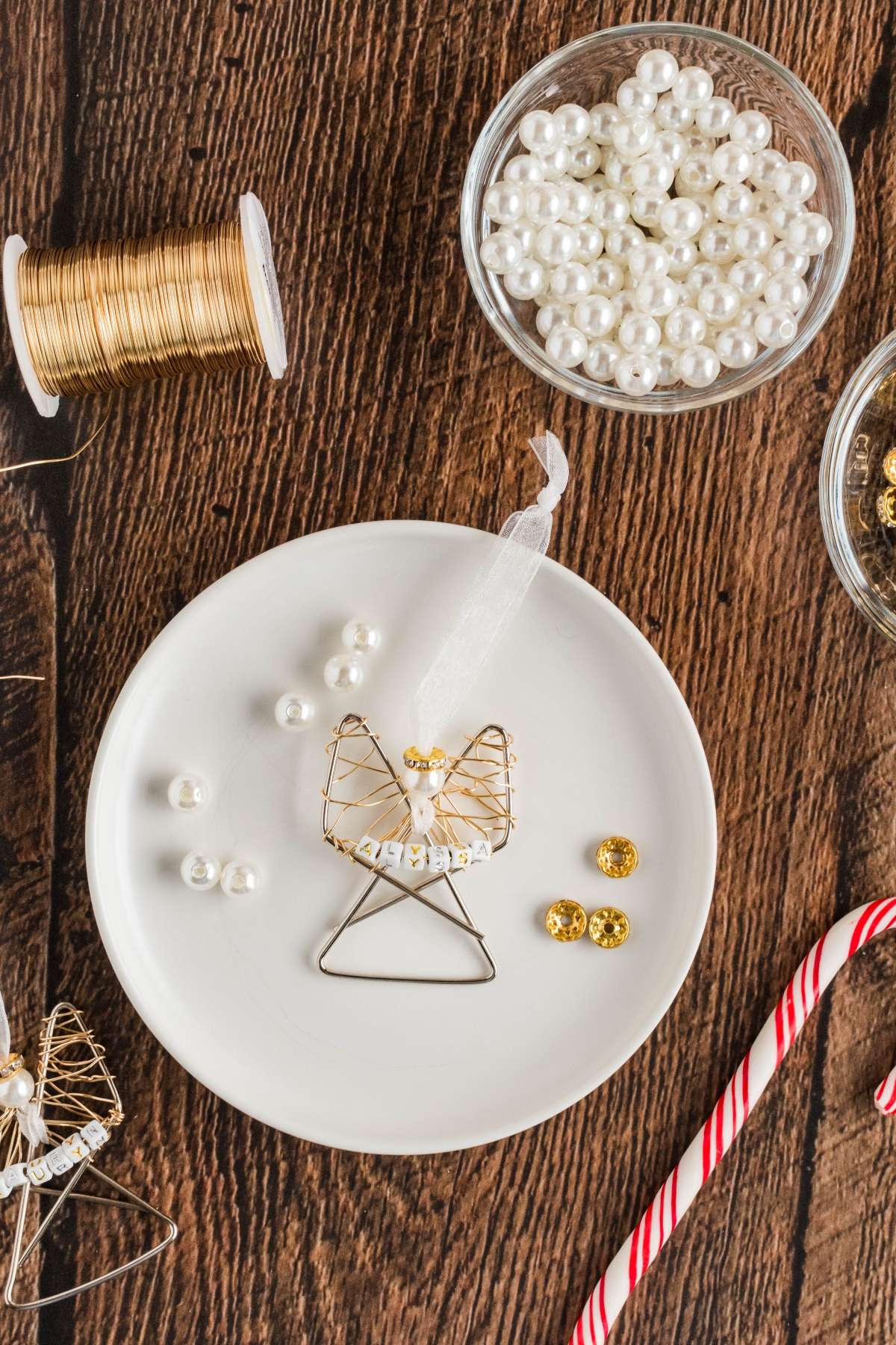 A crafting scene with pearl beads, wire, and a small wire angel on a white plate. A candy cane lies nearby on a wooden surface.